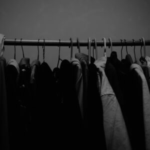 Wieszak z ciasno upchanymi ubraniami w ciemnym świetle – symbol przepełnionej szafy, z której trzeba coś wyrzucić, by zrobić miejsce na nowe podejście.